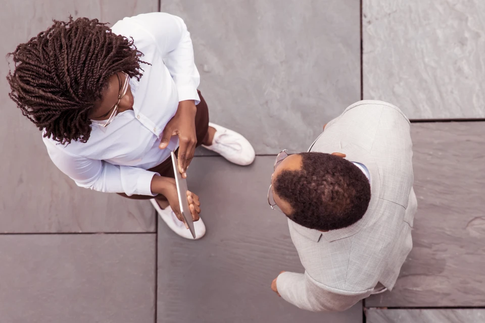 Personne tenant une tablette tactile affichant un canapé gris avec coussins orange en réalité augmentée dans un intérieur domestique minimaliste aux murs blancs et parquet bois clair
